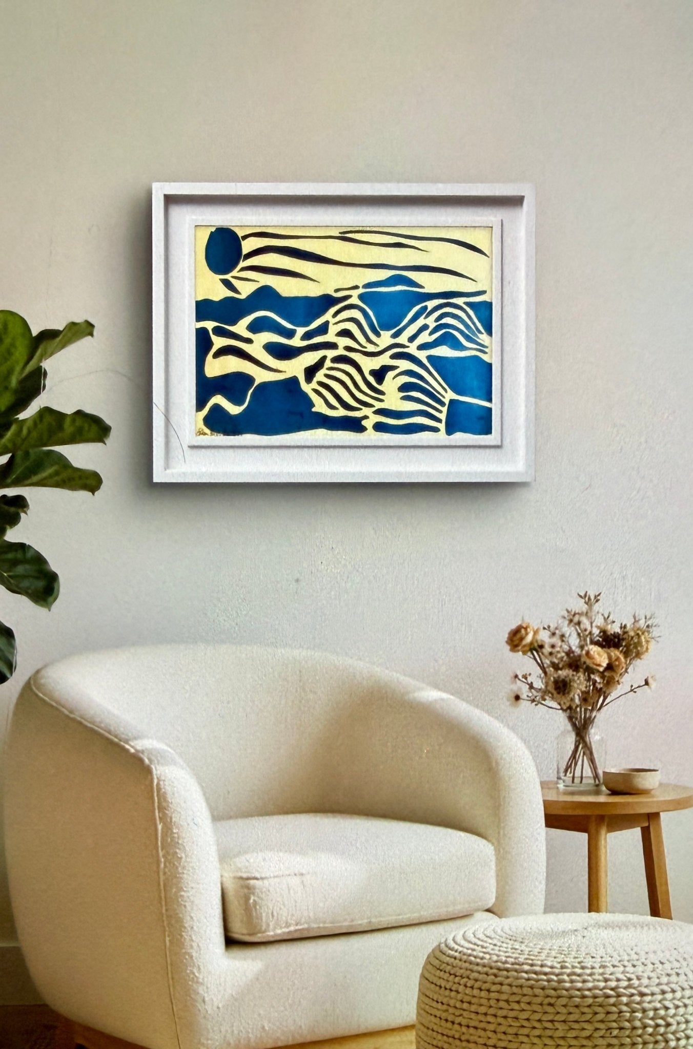 Framed abstract artwork on a wall above a white armchair in a room.