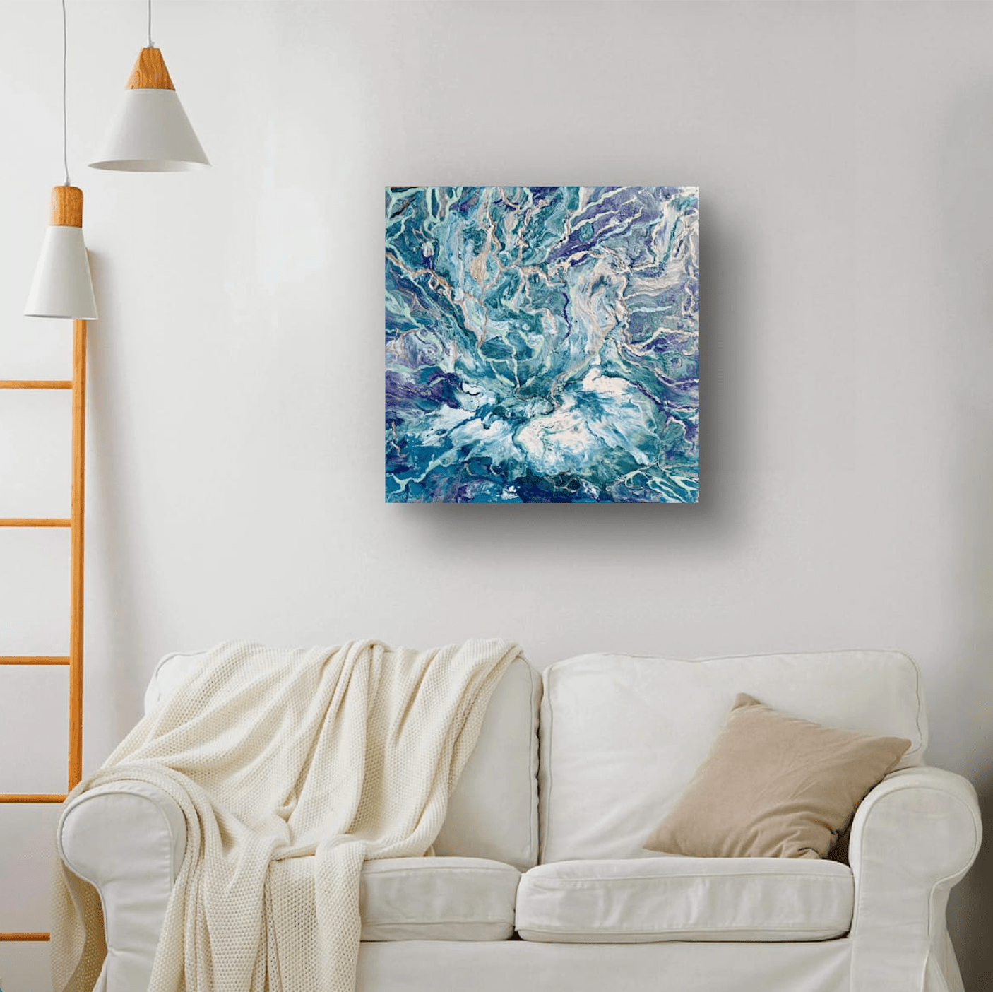 An abstract square canvas print featuring swirling blue, teal, and purple paint patterns, displayed on a white wall above a white sofa with a cream throw blanket.