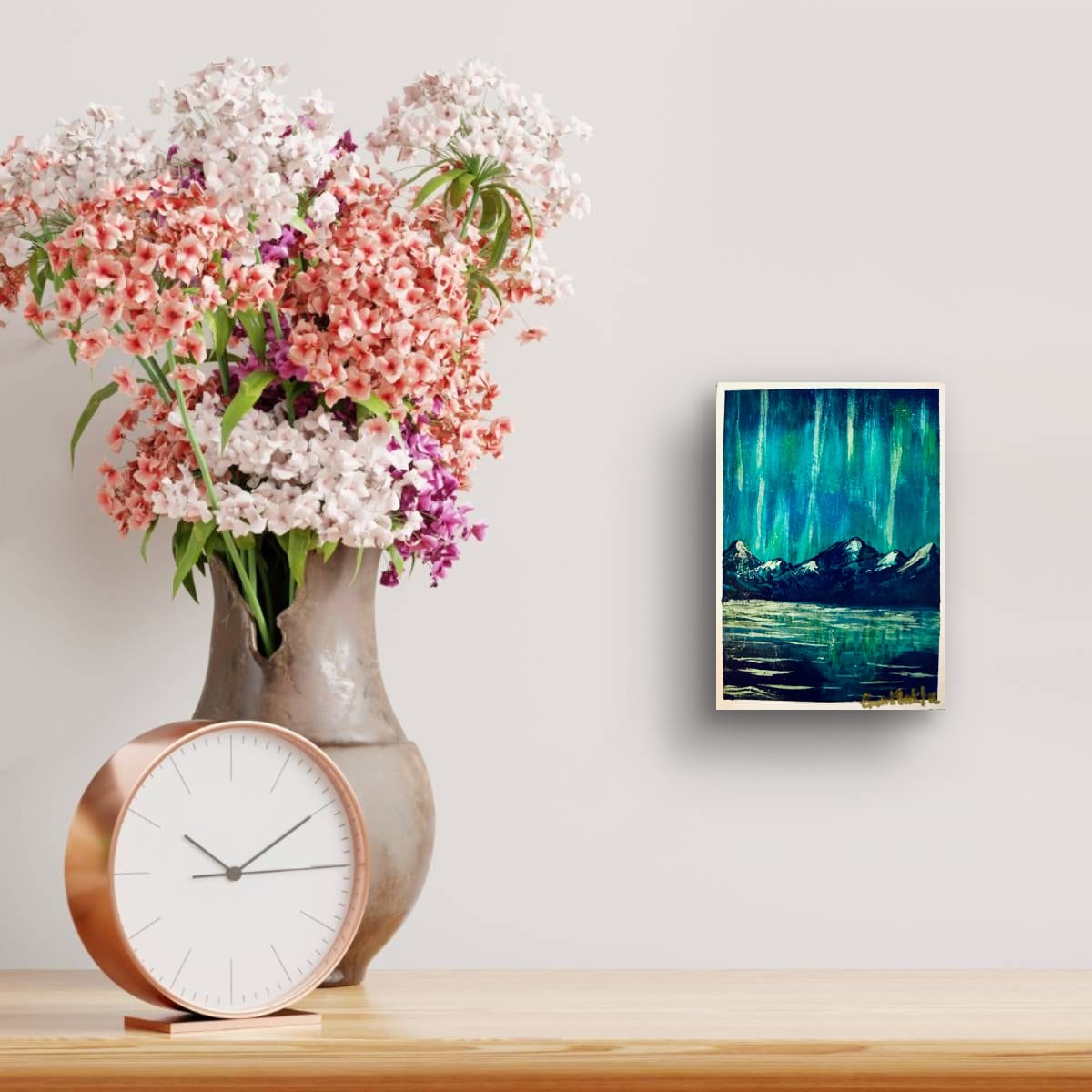Decorative setup with a vase of flowers, clock, and wall art on a light gray wall with an A6 original acrylic mini painting of teal aurora lights over snow-capped mountains with reflections on water by artist Eman Khalifa