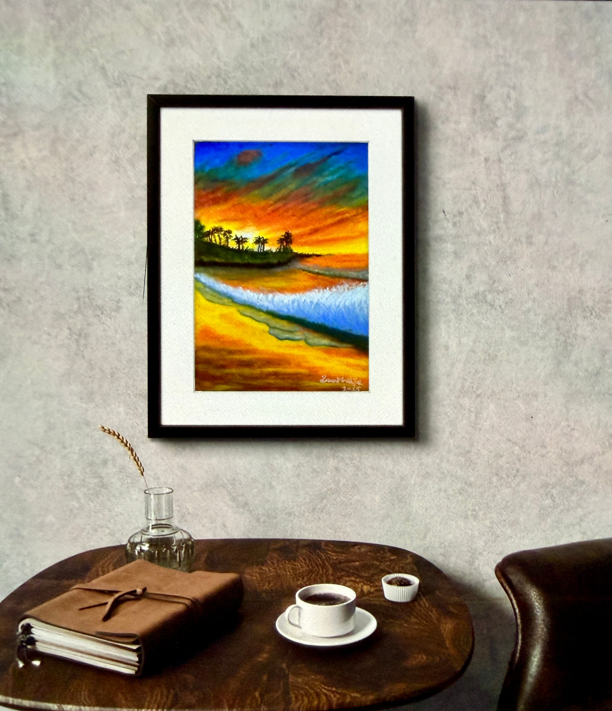 Framed artwork of a beach scene with a firey sunset on a wall above a wooden table with a cup and saucer.