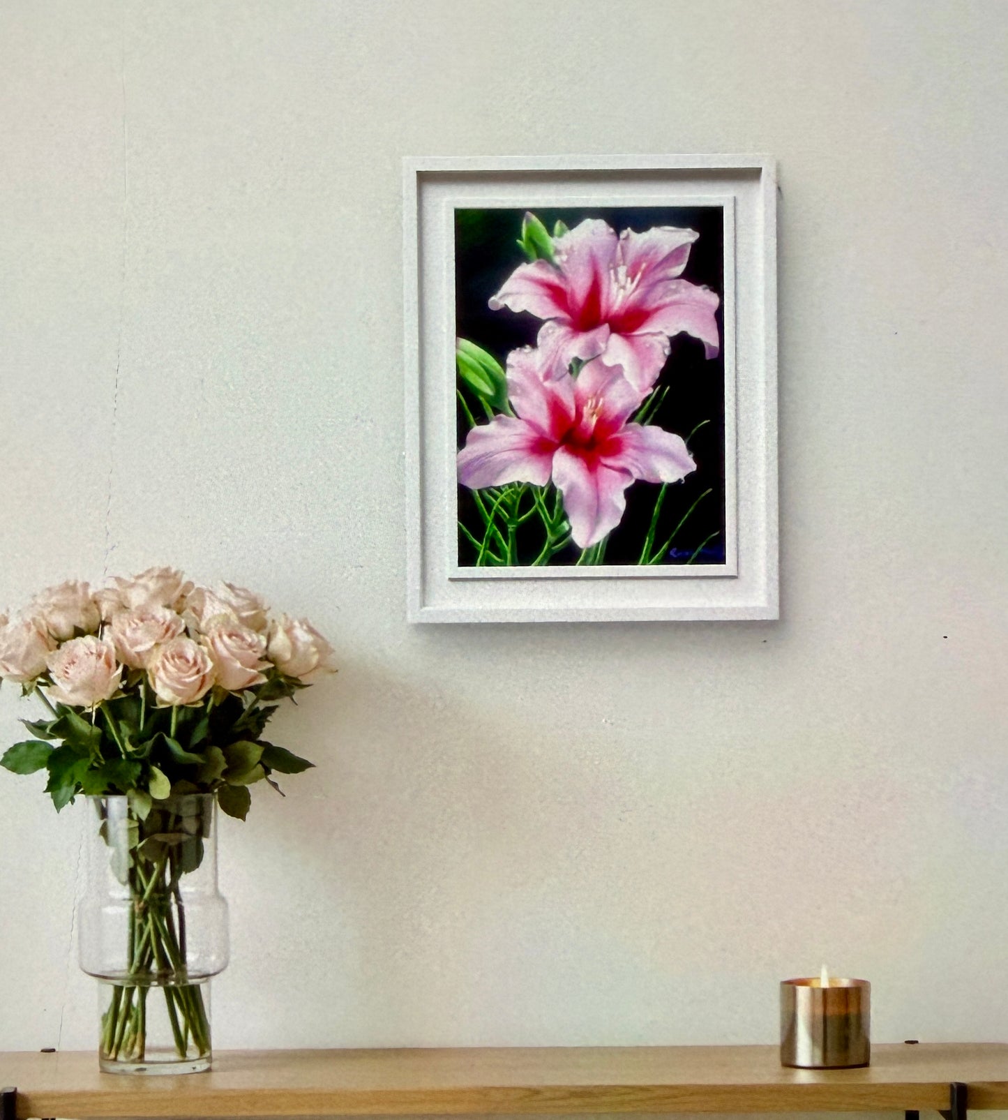 Framed artwork of pink flowers on a wall above a table with flowers and a candle.
