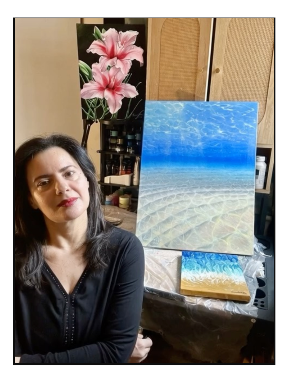 Woman Eman Khalifa sitting next to a painting of a seascape underwater scene and under a pink lilies floral painting