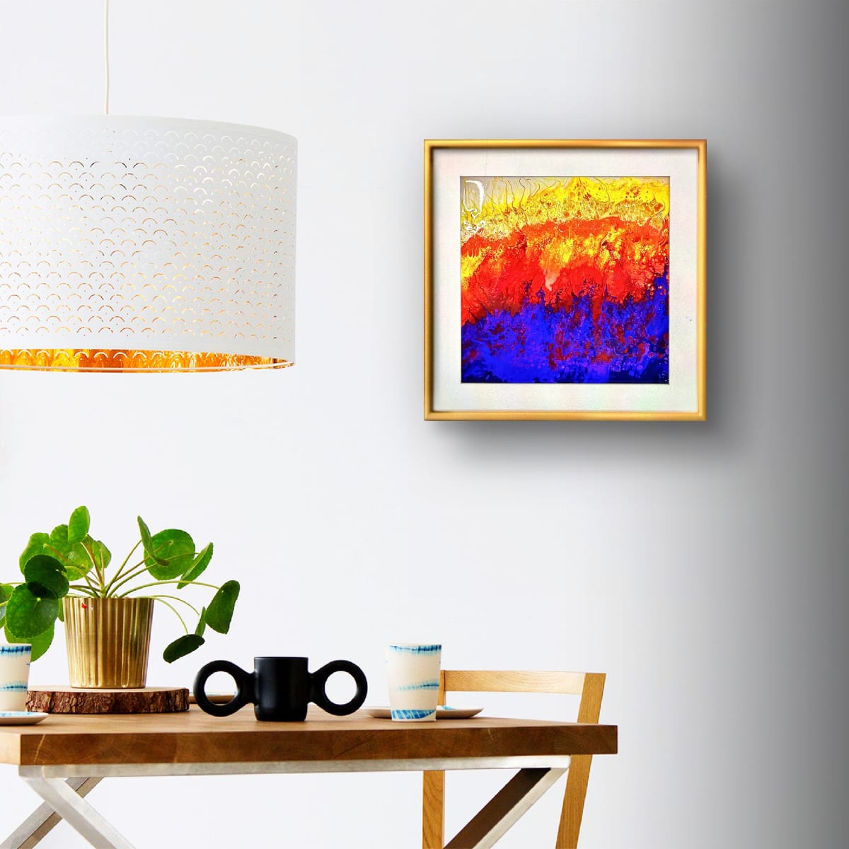 A framed abstract painting with vibrant red, yellow, and blue brushstrokes hangs on a light gray wall above a wooden table styled with a potted plant, a black mug, and a small cup.