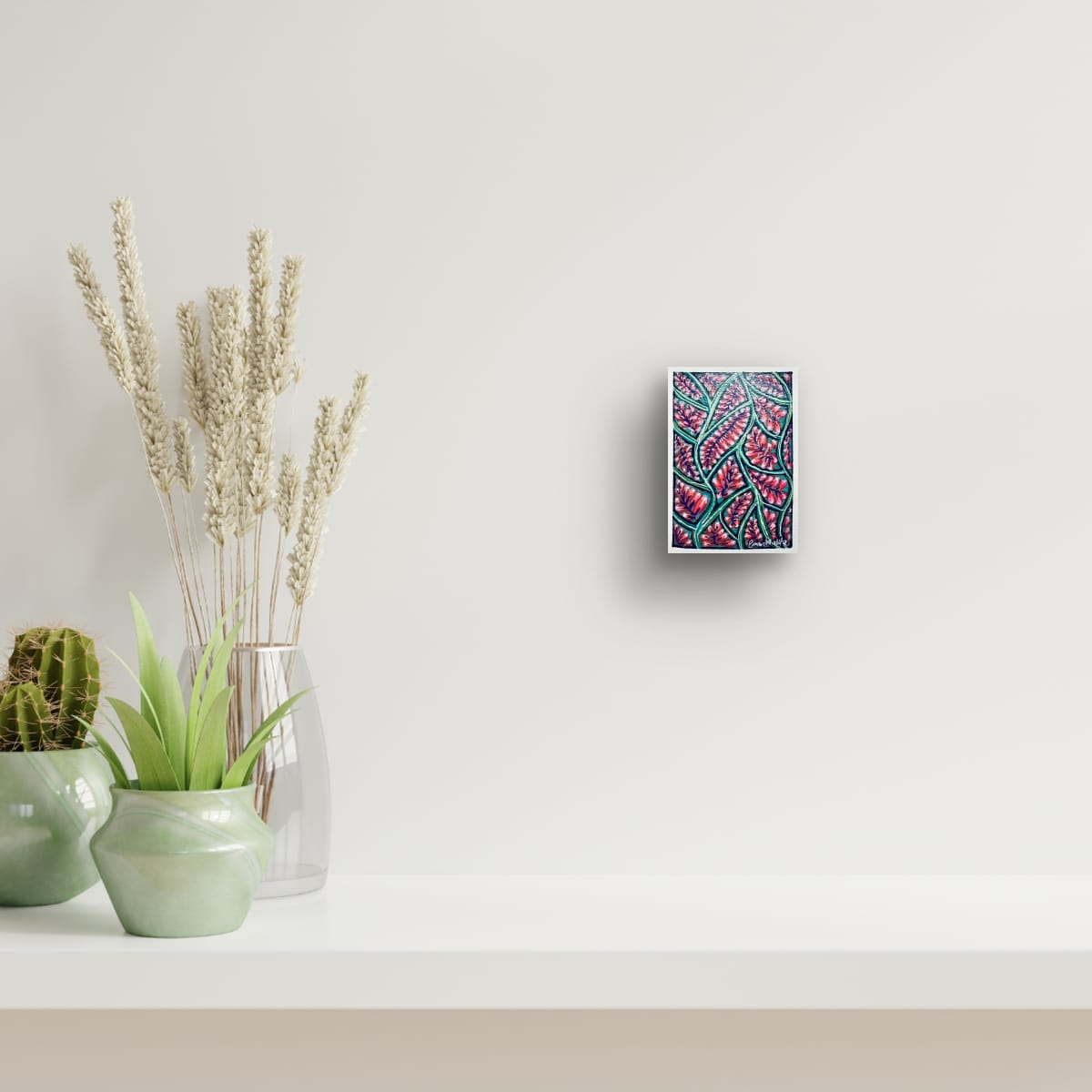 Decorative wall art piece of an A6 original acrylic painting featuring a vibrant floral bloom pattern with red blossoms and green botanical lines by Eman Khalifa, on a white wall with plants below.