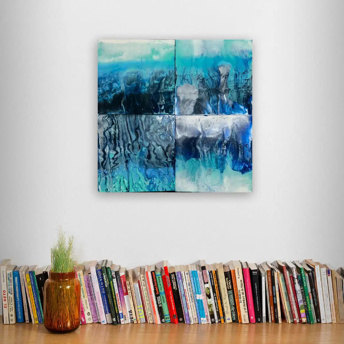 Abstract blue artwork on a wall above a row of books with a vase containing plants.