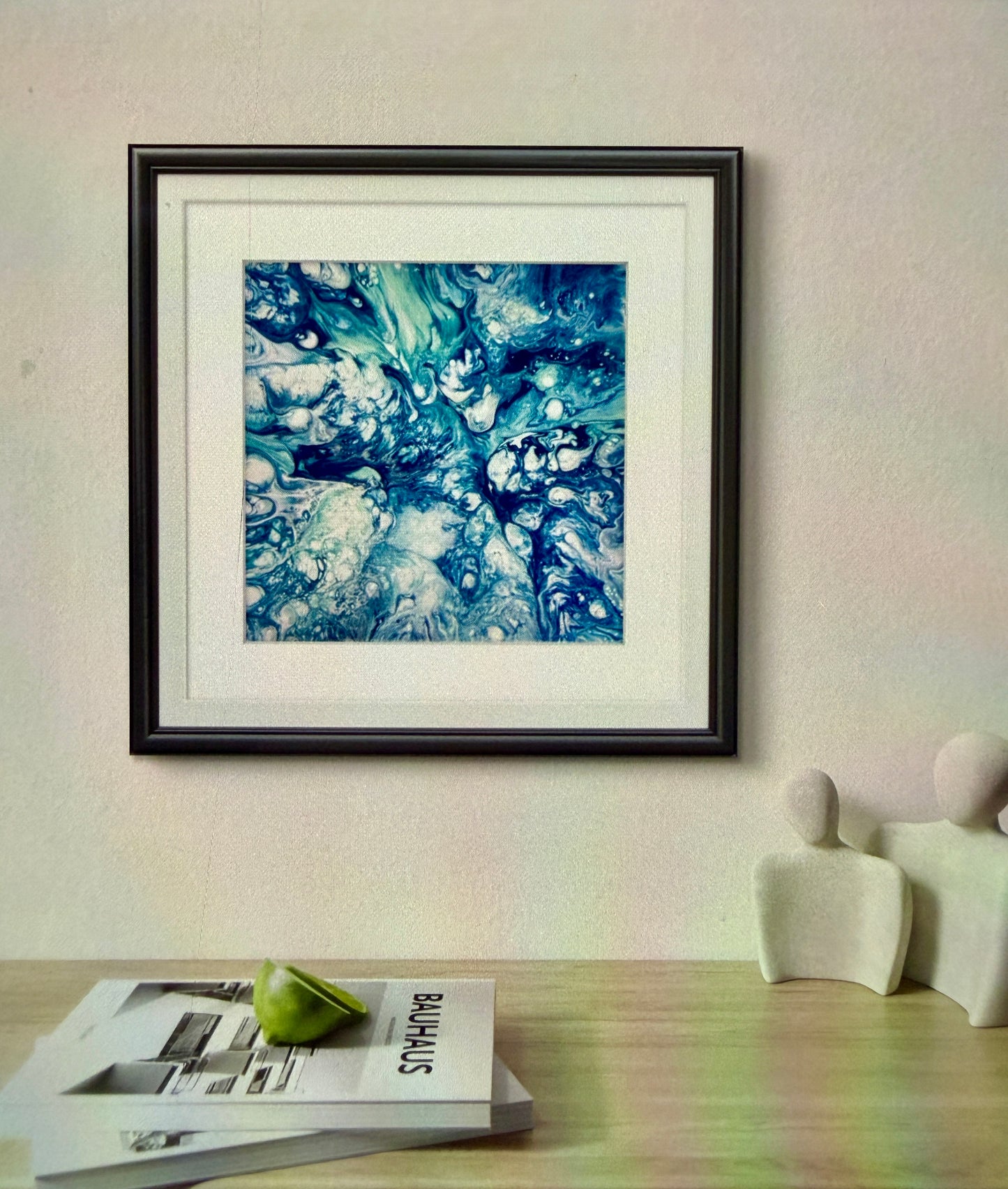 Framed abstract art piece on a wall with a table and books in the foreground.