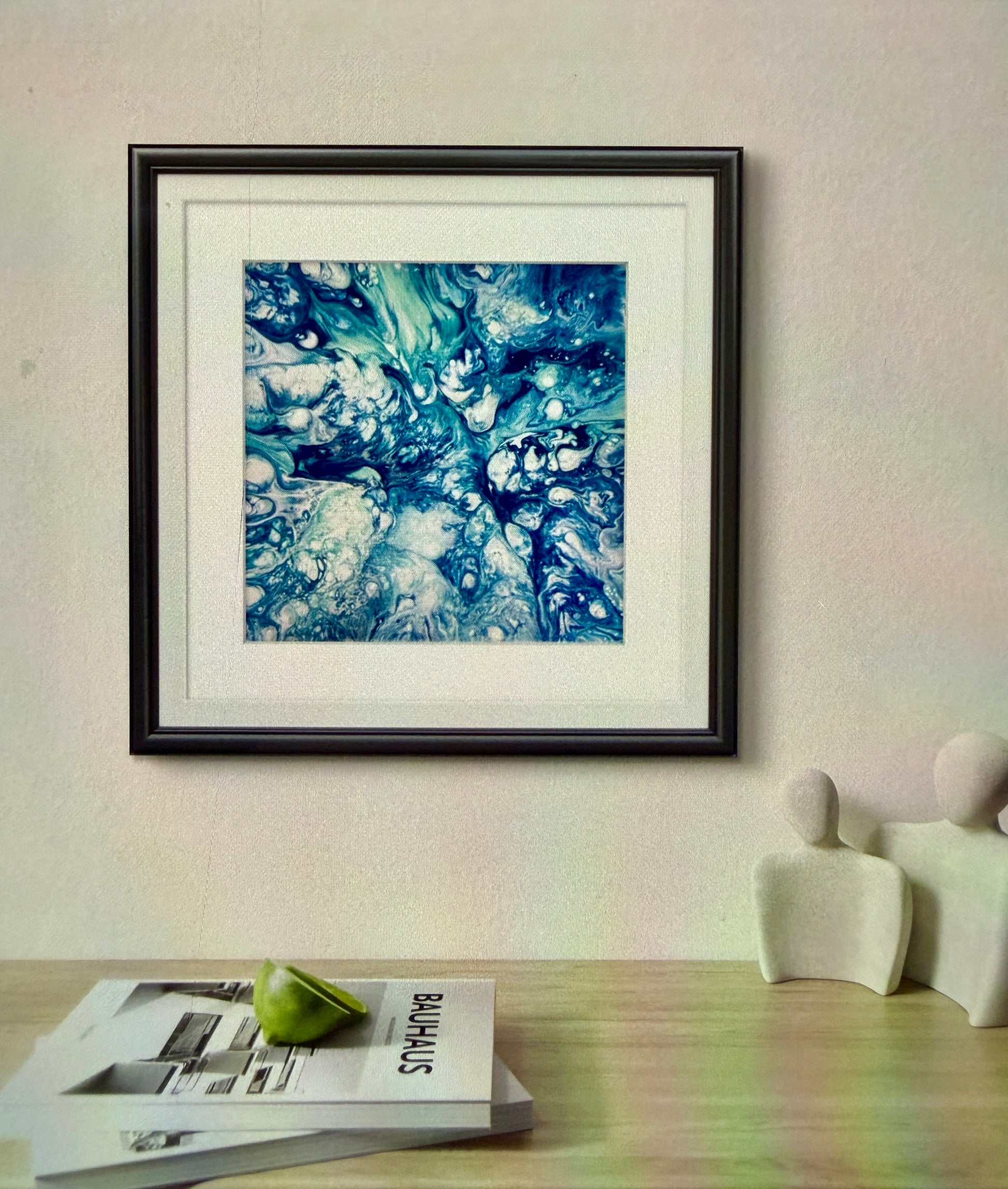 Framed abstract art piece on a wall with a table and books in the foreground.