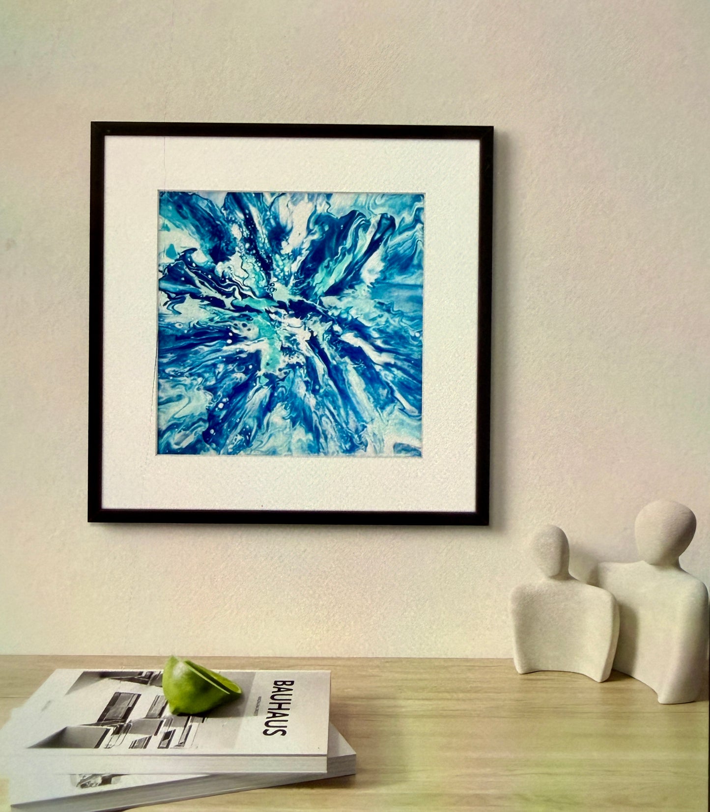 Framed abstract blue artwork on a wall above a table with magazines and a decorative item.