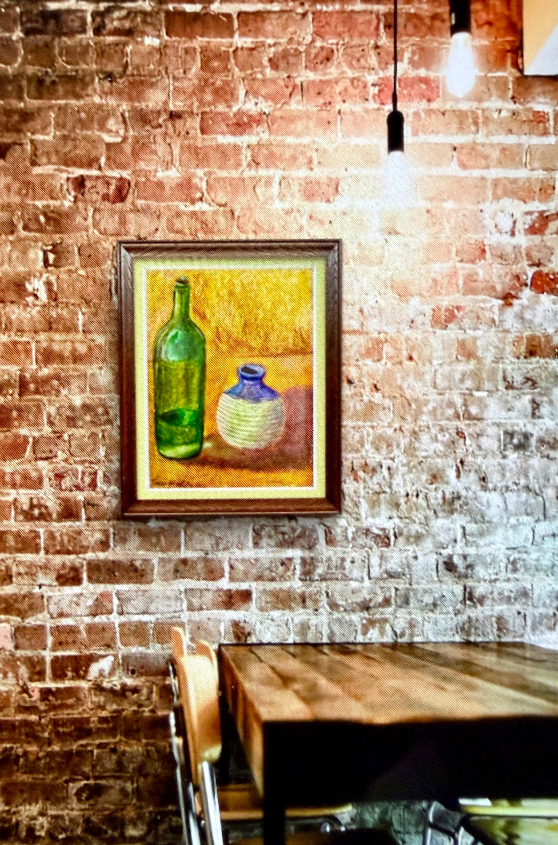 Framed painting of bottles on a brick wall with a wooden table below.