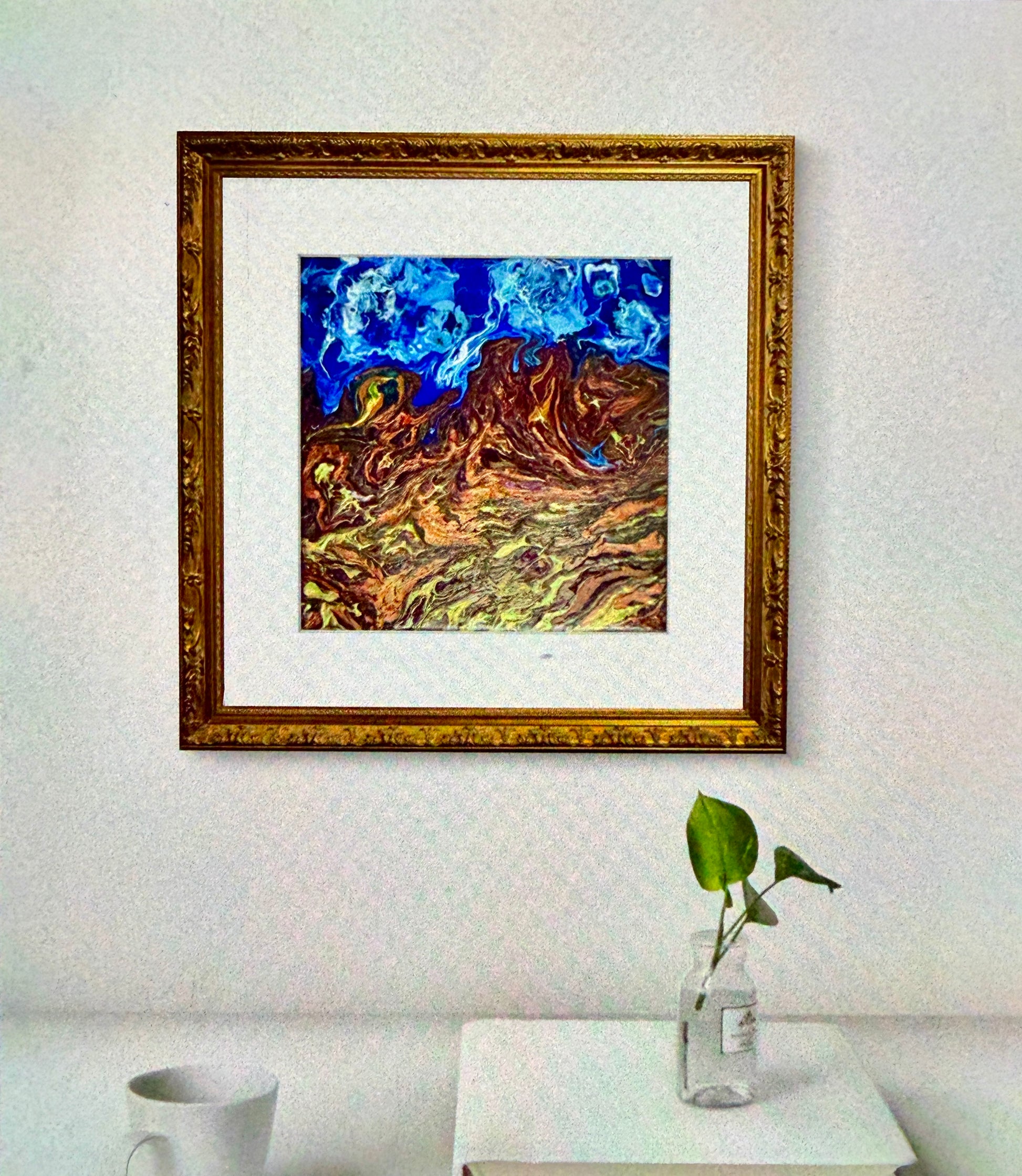 Framed abstract painting on a wall with a plant and cup on a table below.