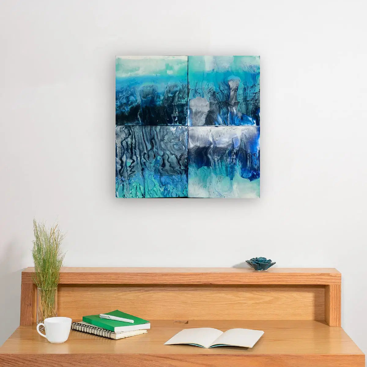 Abstract blue artwork on a white wall above a wooden desk with books and a cup.